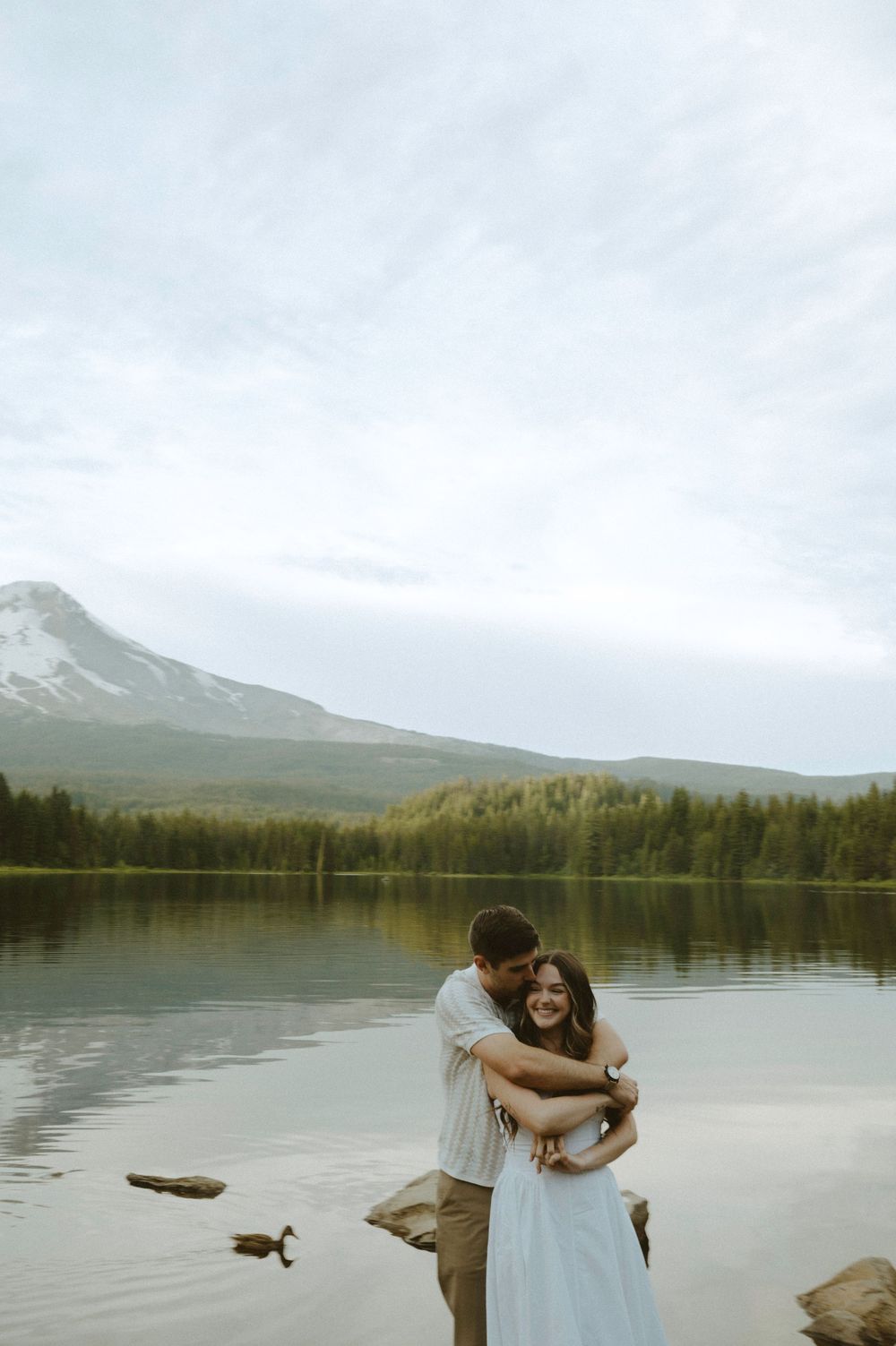 Couple embracing and smiling by a serene mountain lake with evergreen trees and a snow-capped peak in the background.