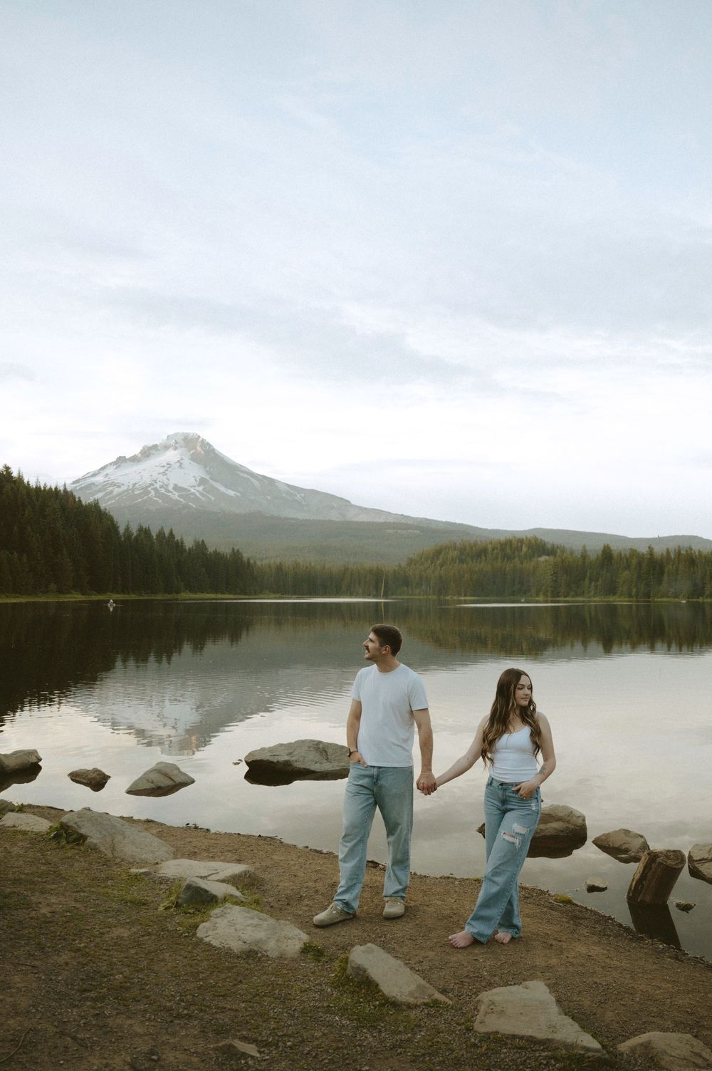 Couple holding hands by a serene lake with Mount Hood reflected in the water, surrounded by evergreen trees.