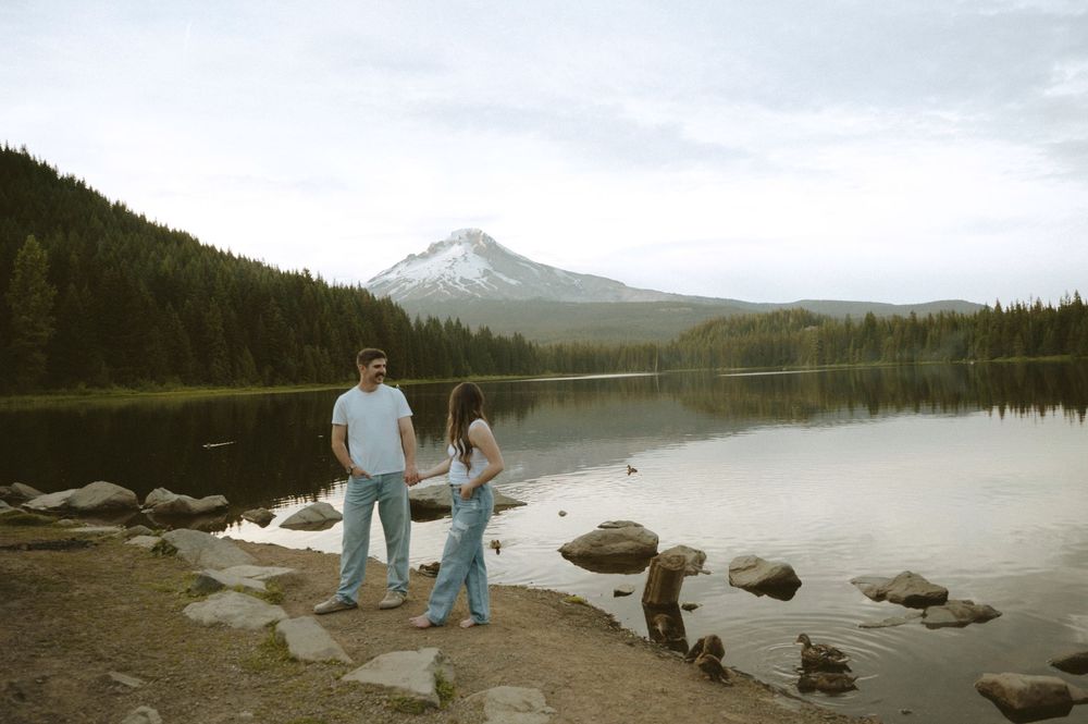 Couple holding hands by a serene mountain lake with forest and snow-capped peak reflected in calm water.