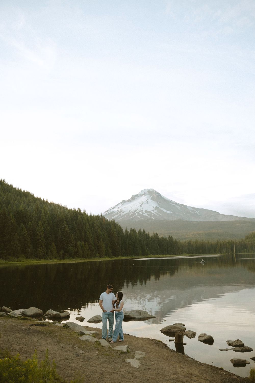 Couple standing together at Trillium Lake shore with snow-capped Mount Hood and forest reflection in calm water.