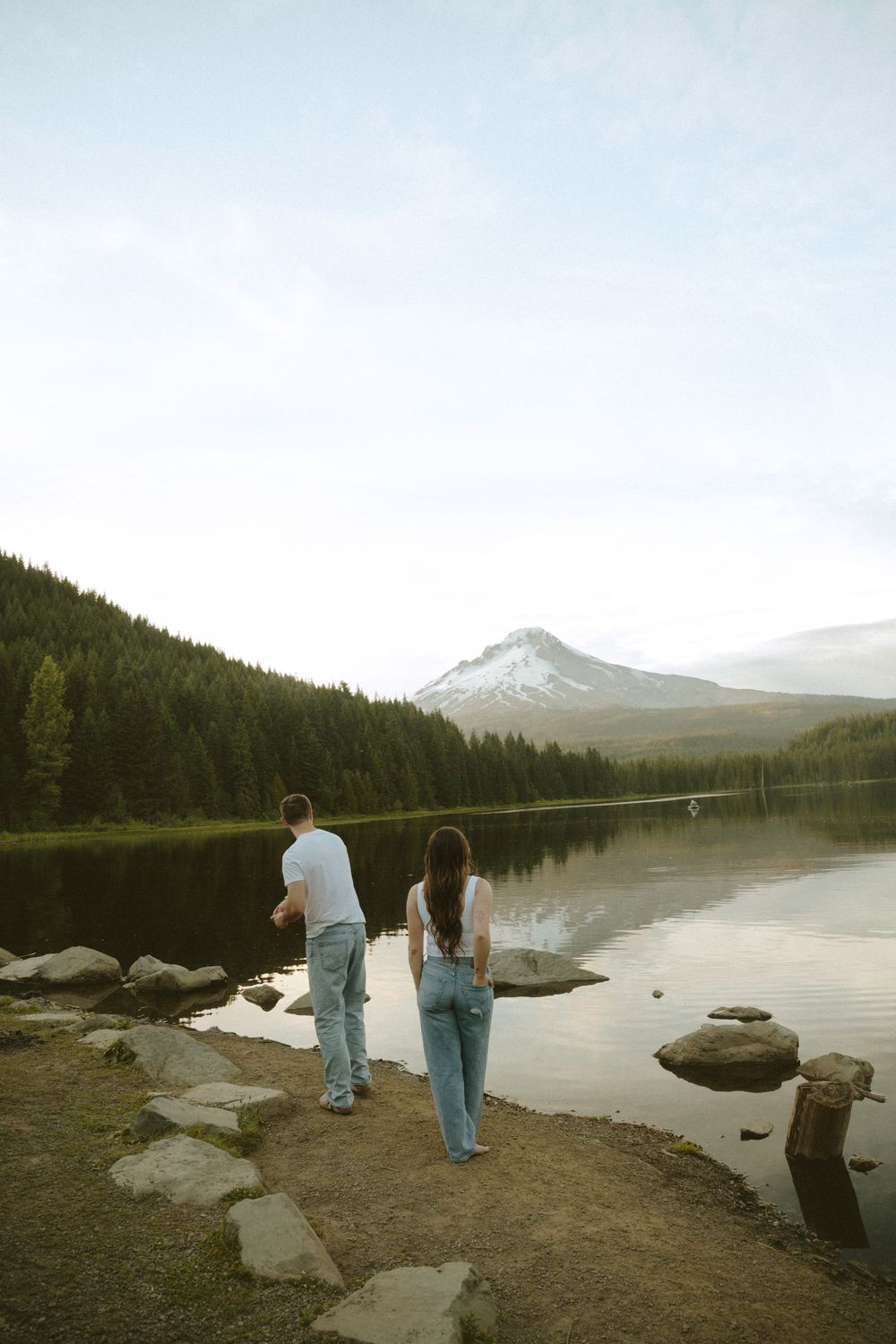 Couple standing by a serene mountain lake with Mt. Hood reflection, surrounded by evergreen forest in Oregon.