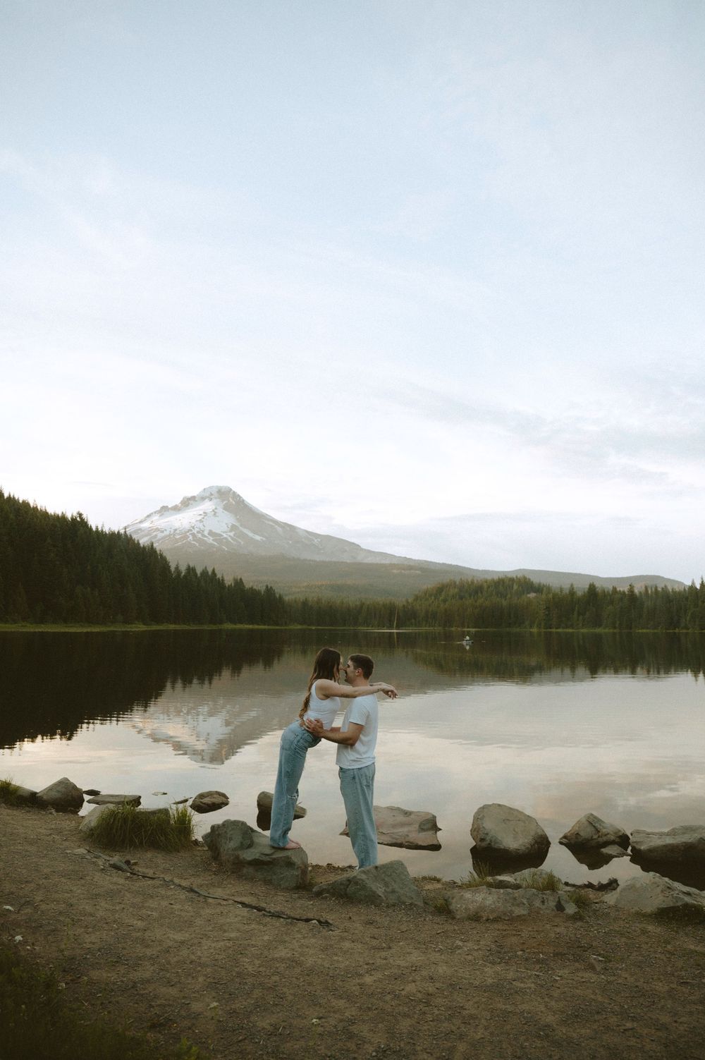 Couple embracing by a serene mountain lake with Mt. Hood reflection, surrounded by evergreen trees and rocky shoreline.