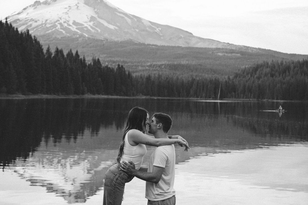 Couple kissing by a serene lake with a snow-capped mountain and pine forest in black and white.
