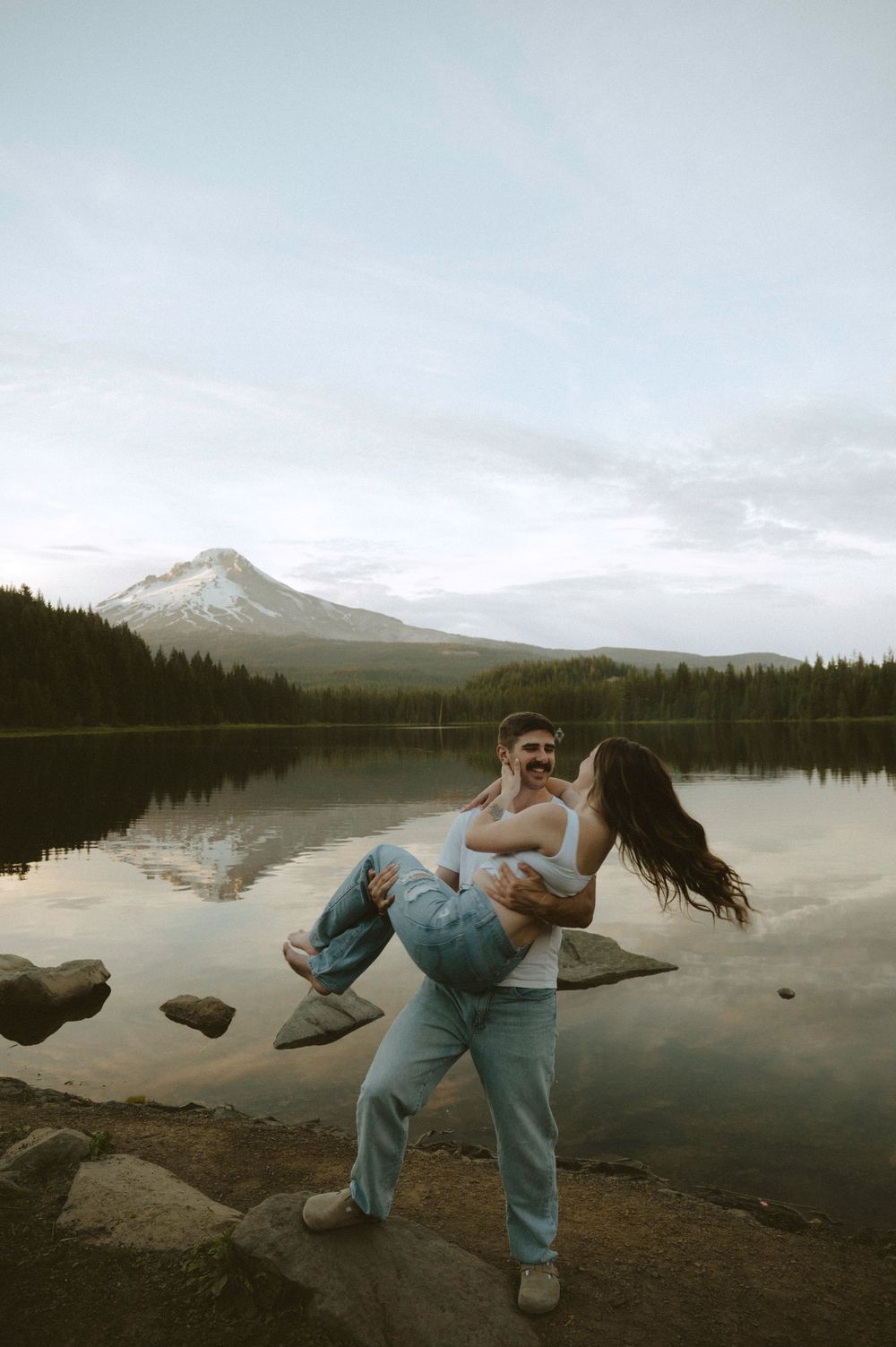 A man lifts a woman in his arms by a serene lake with Mount Hood reflected in the water at dusk.