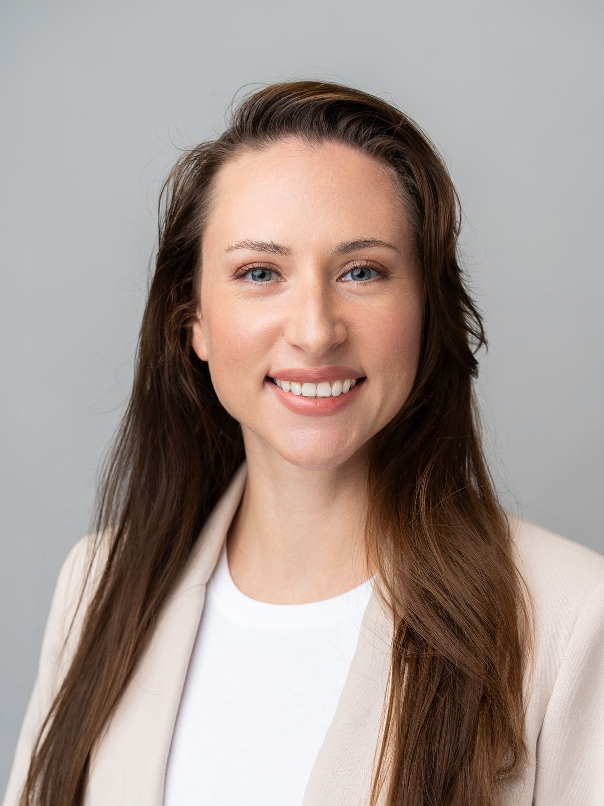 Professional headshot of a smiling young woman with long brown hair wearing a beige blazer and white top.