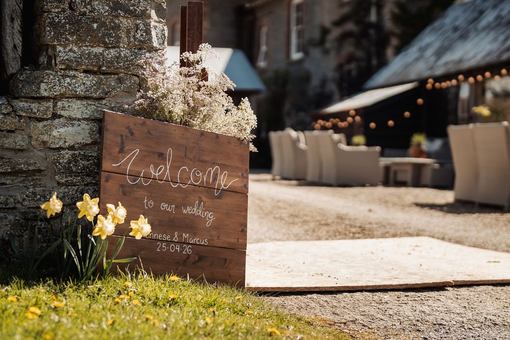 Rustic wooden welcome wedding sign with daffodils beside a stone venue with fairy lights and outdoor seating.
