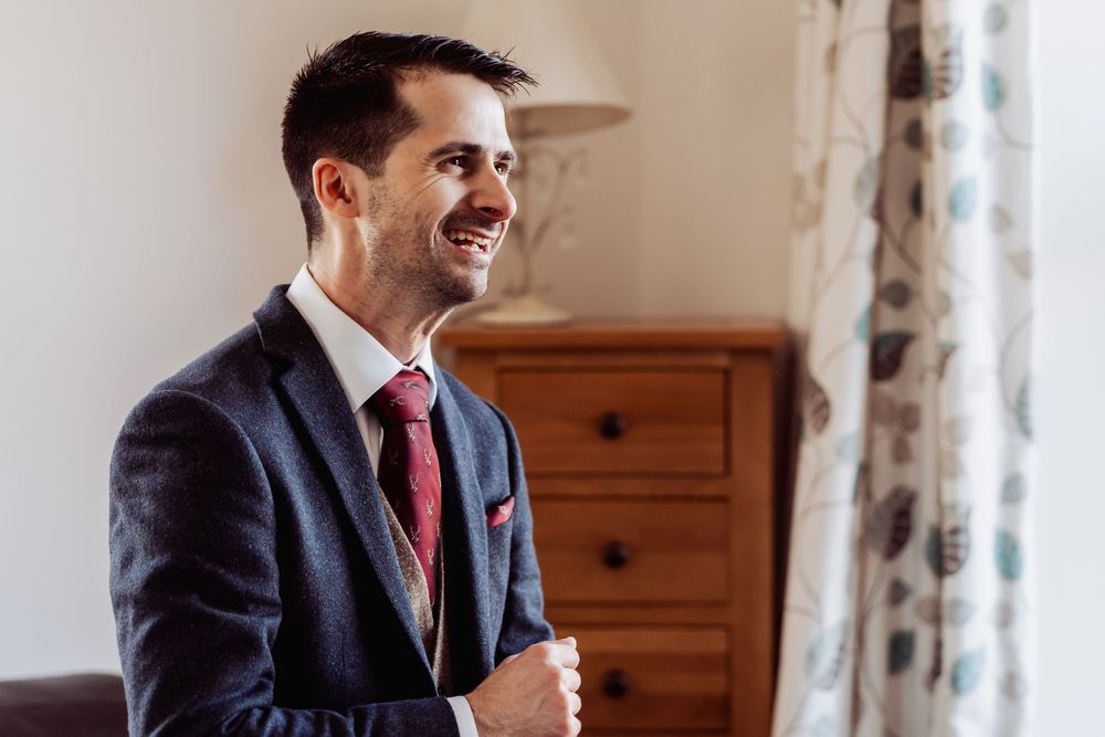 Smiling groom in navy suit and red tie adjusting jacket near window on wedding day.