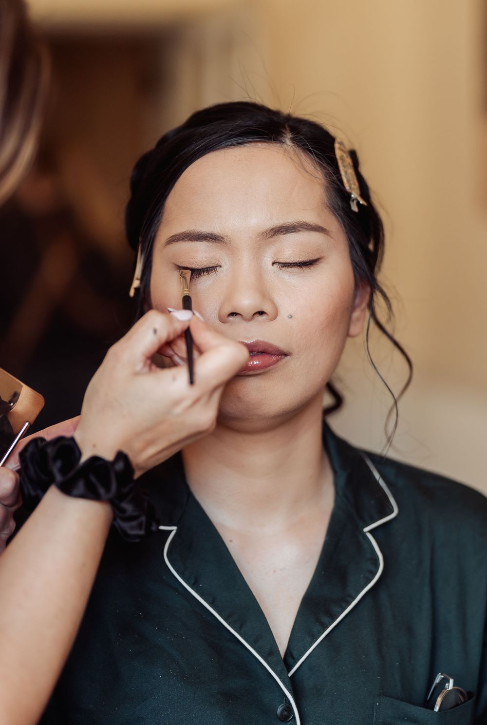 A makeup artist applies eyeliner to an Asian woman in a green satin robe, with hair clips and a black scrunchie visible.