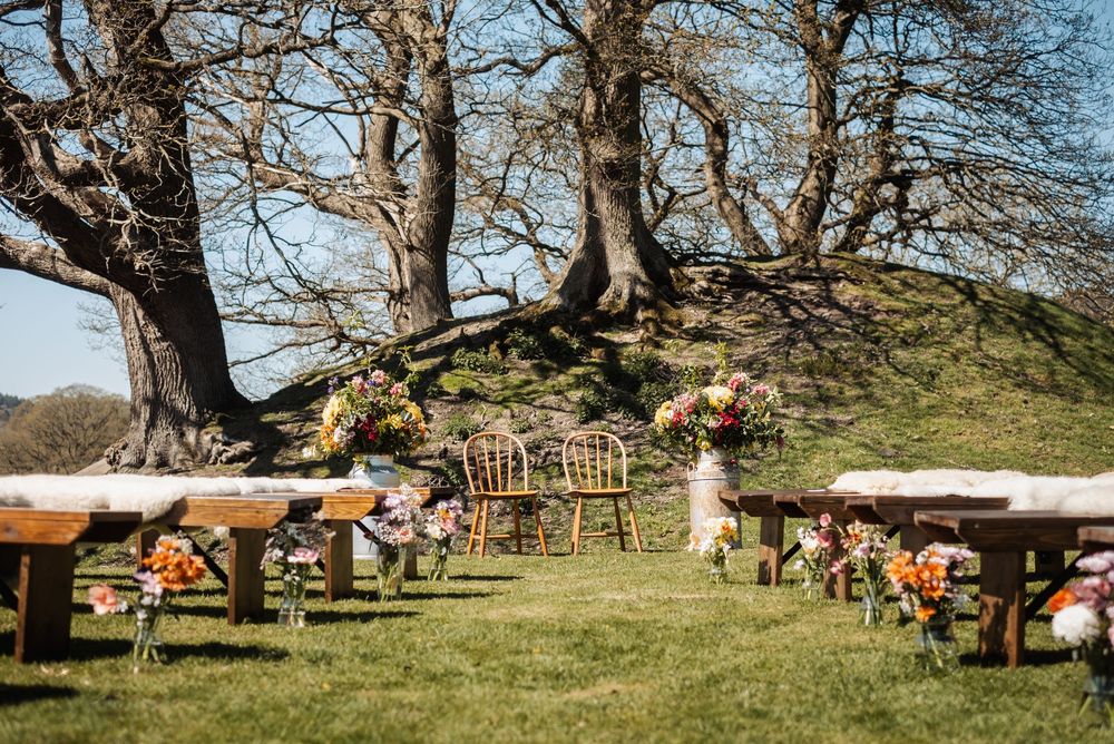 Outdoor wedding ceremony setup with wooden benches, floral arrangements, and two chairs under bare trees on a green lawn.