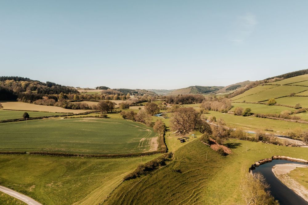 Aerial view of lush green rolling hills and farmland valleys under a clear blue sky in the British countryside.