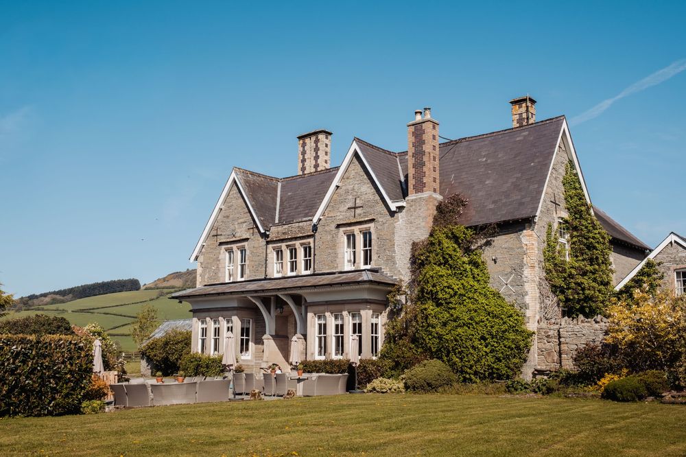 A grand stone country manor house with manicured lawns, ivy-covered walls, and rolling green hills under a clear blue sky.