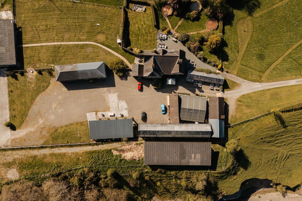 Aerial drone view of a rural farm complex with multiple buildings, gravel paths, and green fields on a sunny day.