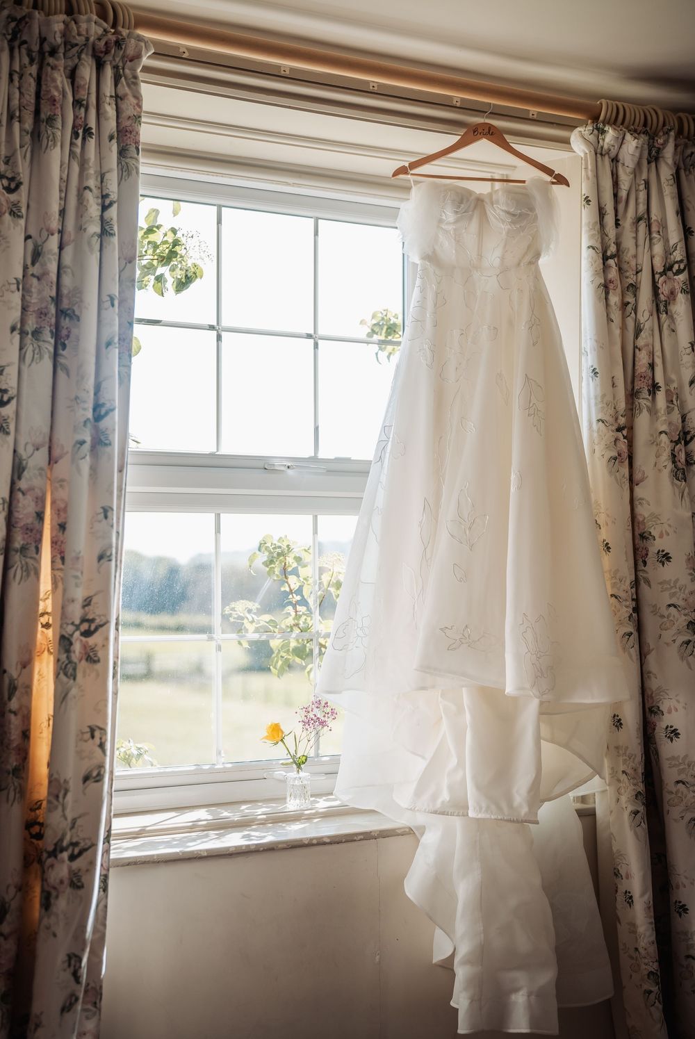 White strapless wedding dress hanging on wooden hanger in sunlit window with floral curtains and small flower vases below.