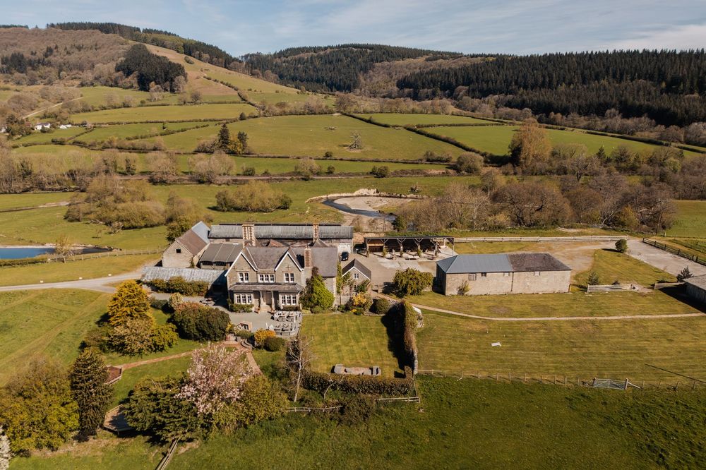 Aerial view of a countryside estate with stone buildings surrounded by lush green fields and wooded hills.