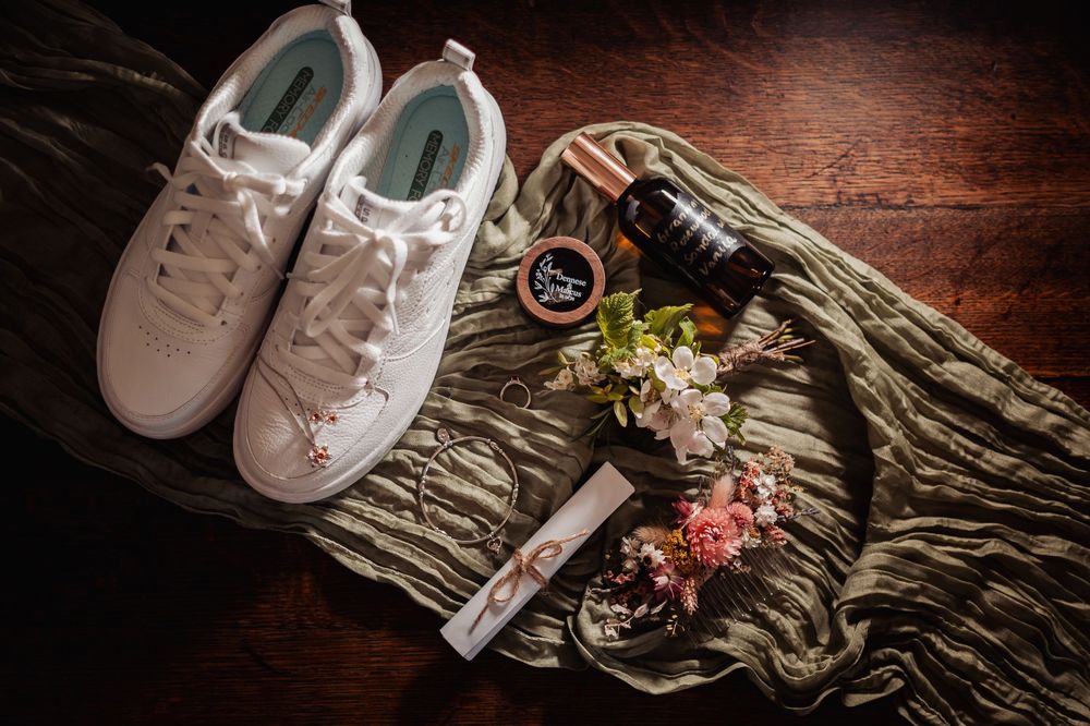 White sneakers with floral accents beside perfume, lip balm, and pink flowers on a green scarf.