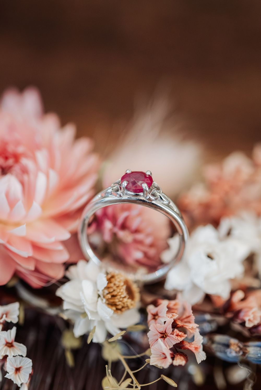 Silver ring with red ruby gemstone surrounded by pink and white flowers on a dark wooden background.