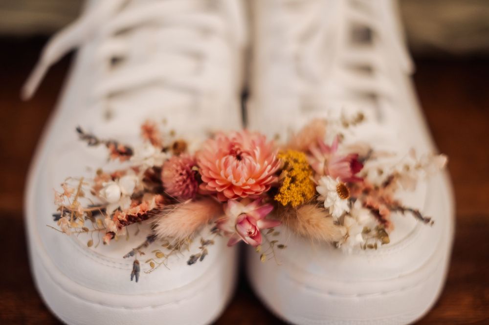White sneakers adorned with dried floral arrangement featuring pink, coral, and mustard flowers on wooden surface.