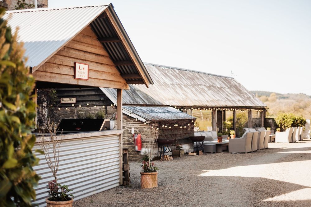 Rustic outdoor venue with wooden barn structures, string lights, gravel courtyard, and cozy lounge seating surrounded by lush greenery.