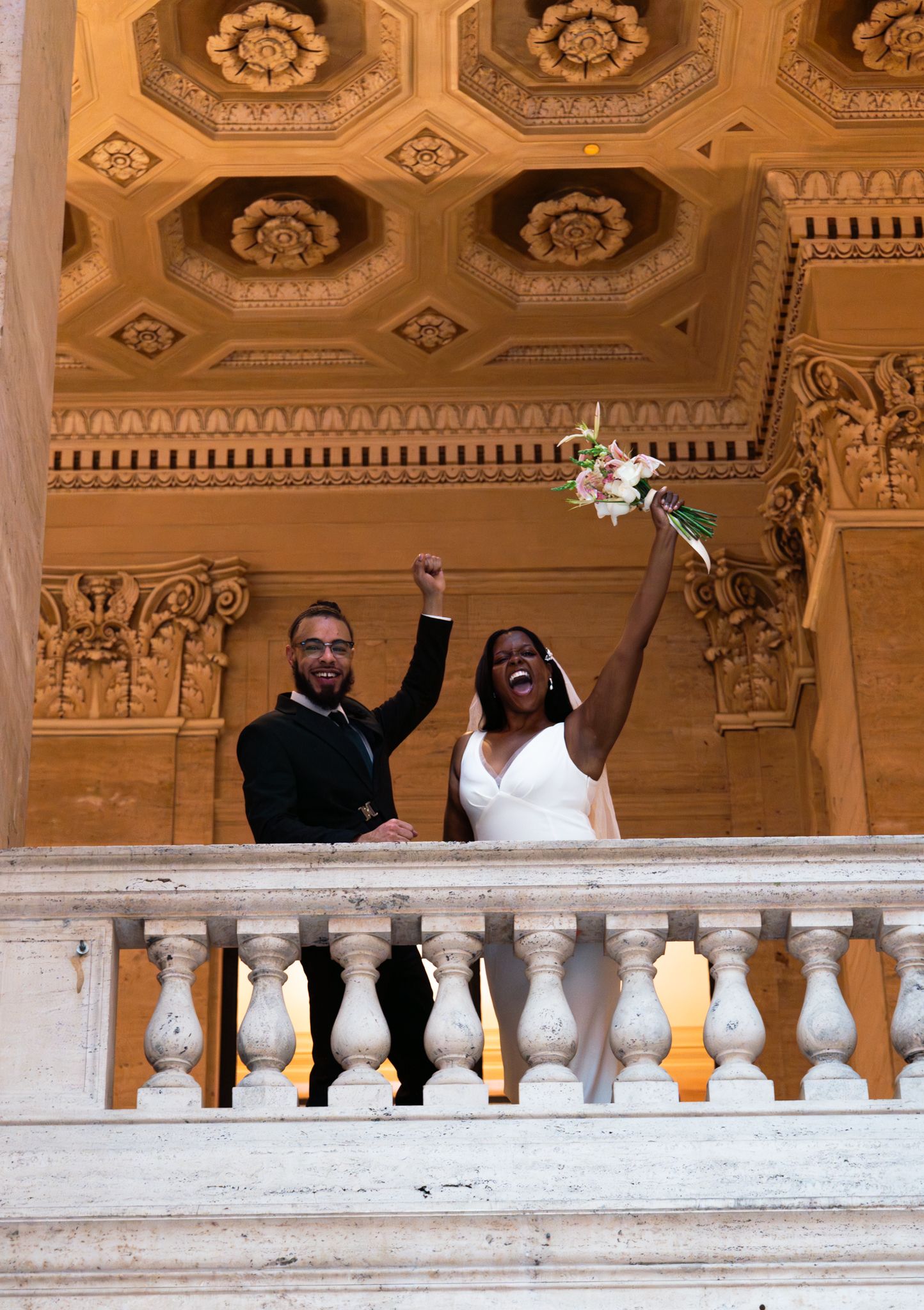 Joyful newlyweds celebrate on ornate marble balcony, bride raising bouquet and groom pumping fist in elegant gold-ceilinged venue.