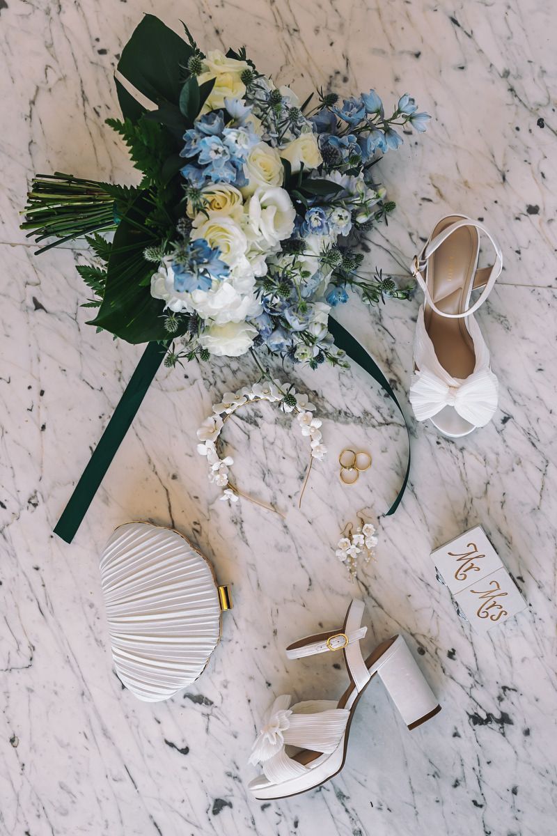 Bridal flat lay with blue and white bouquet, white heels, clutch purse, floral crown, and gold rings on marble.