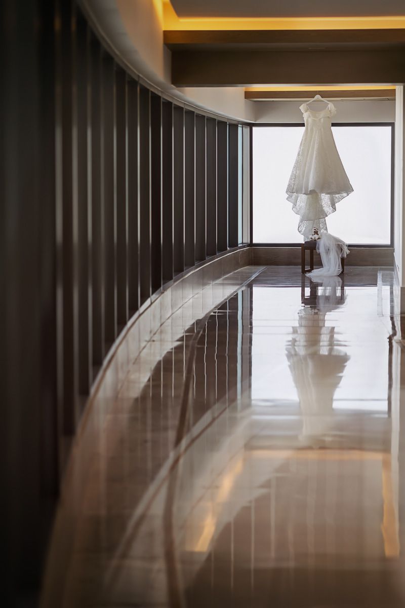 Elegant white wedding dress hanging near a large window in a luxurious hotel corridor with marble floors.