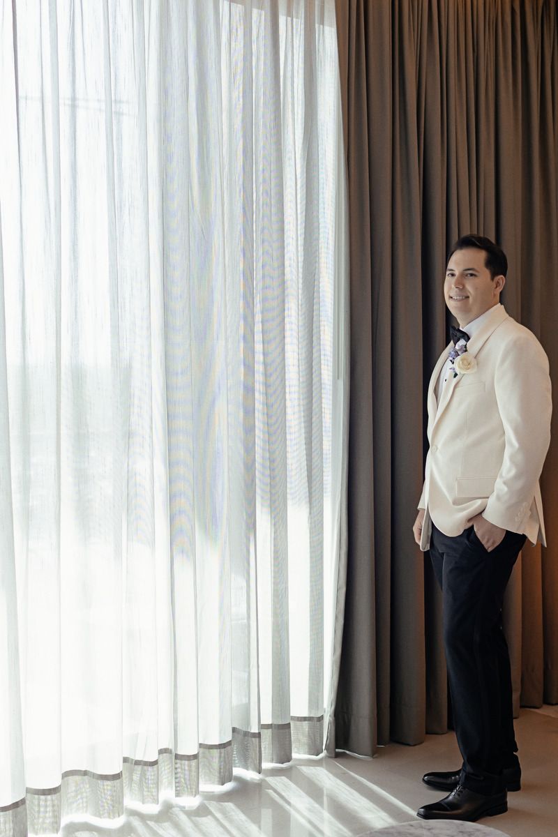 Groom in white jacket and black pants standing by sheer curtains near a bright window on wedding day.