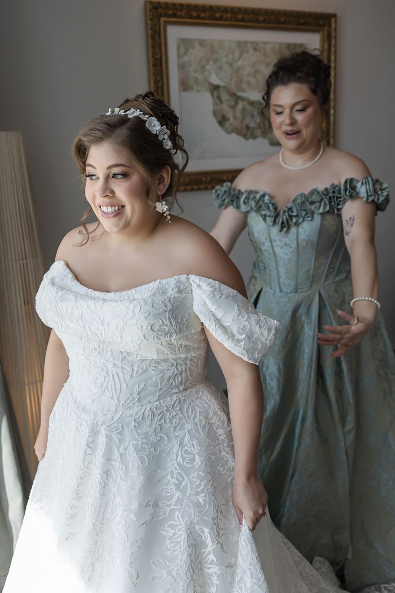 Bride in off-shoulder lace gown smiles as bridesmaid in teal corset dress helps her prepare for wedding.