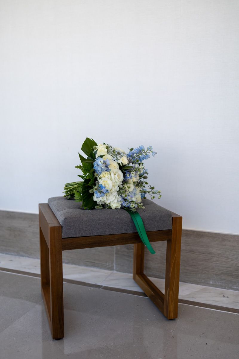 A bridal bouquet of white and blue flowers with a green ribbon rests on a wooden stool with gray upholstery.