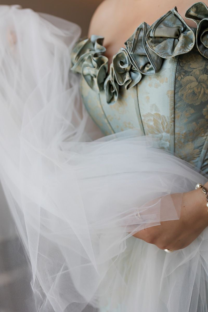 Close-up of bride in teal floral corset gown with ruffled details holding flowing white tulle skirt.