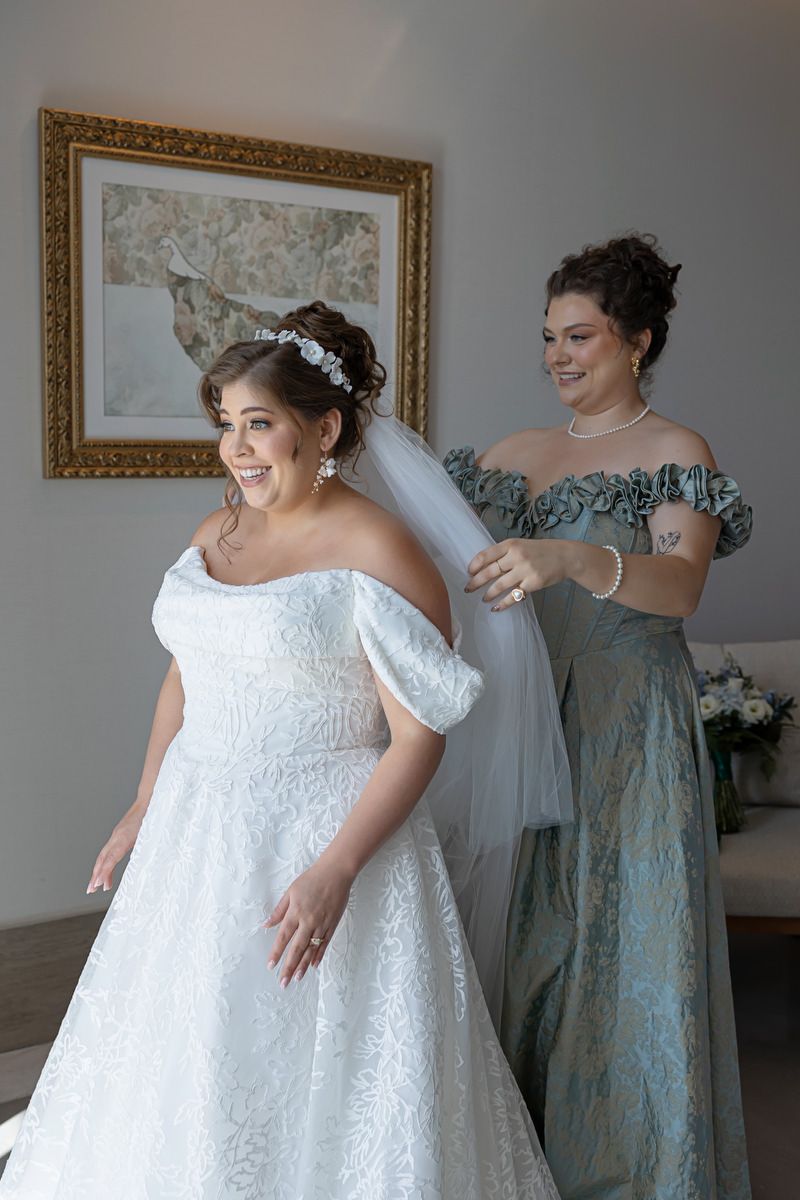 Bride in white lace gown smiles as bridesmaid in sage green dress adjusts her veil before the wedding ceremony.