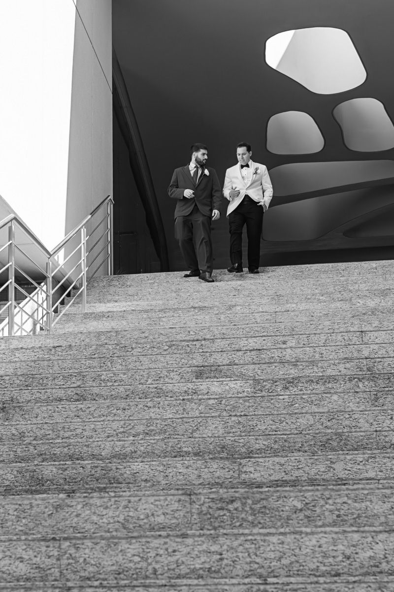 Black and white photo of two groomsmen in suits walking down outdoor stone steps at wedding venue.