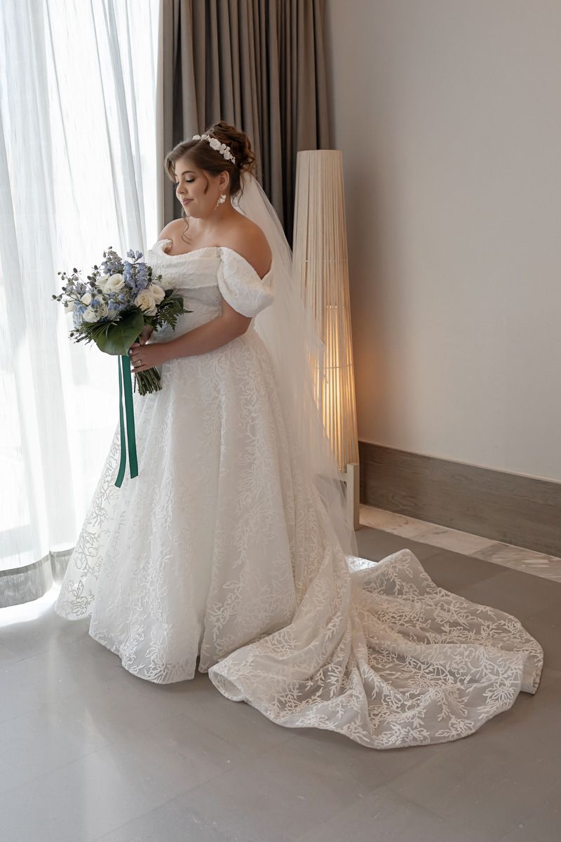 Bride in off-shoulder lace wedding gown with cathedral train holds blue and white bouquet by a sunlit window.