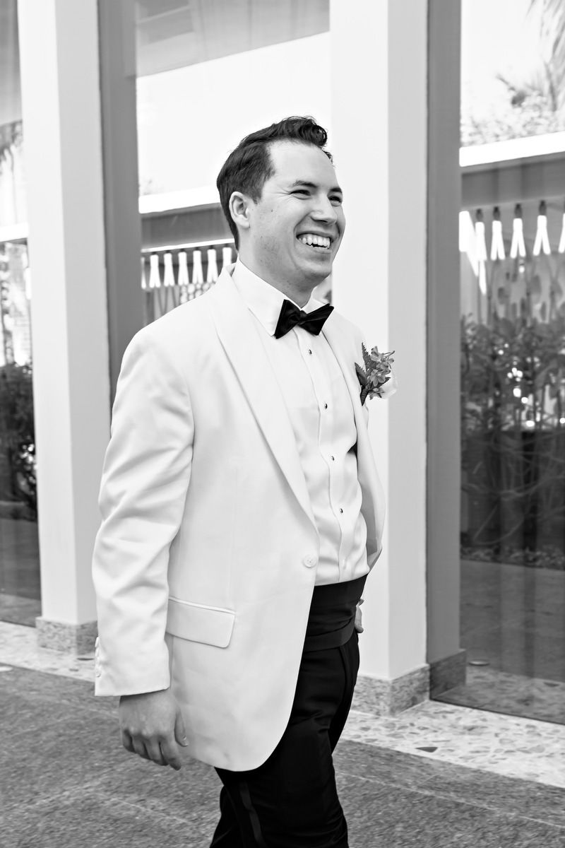 A smiling groom in a white tuxedo jacket with black bow tie and boutonniere walks outdoors in black and white photo.
