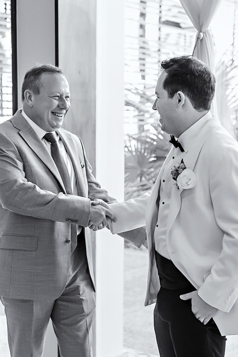 Groom in white tuxedo shakes hands with smiling older man in gray suit at wedding venue.