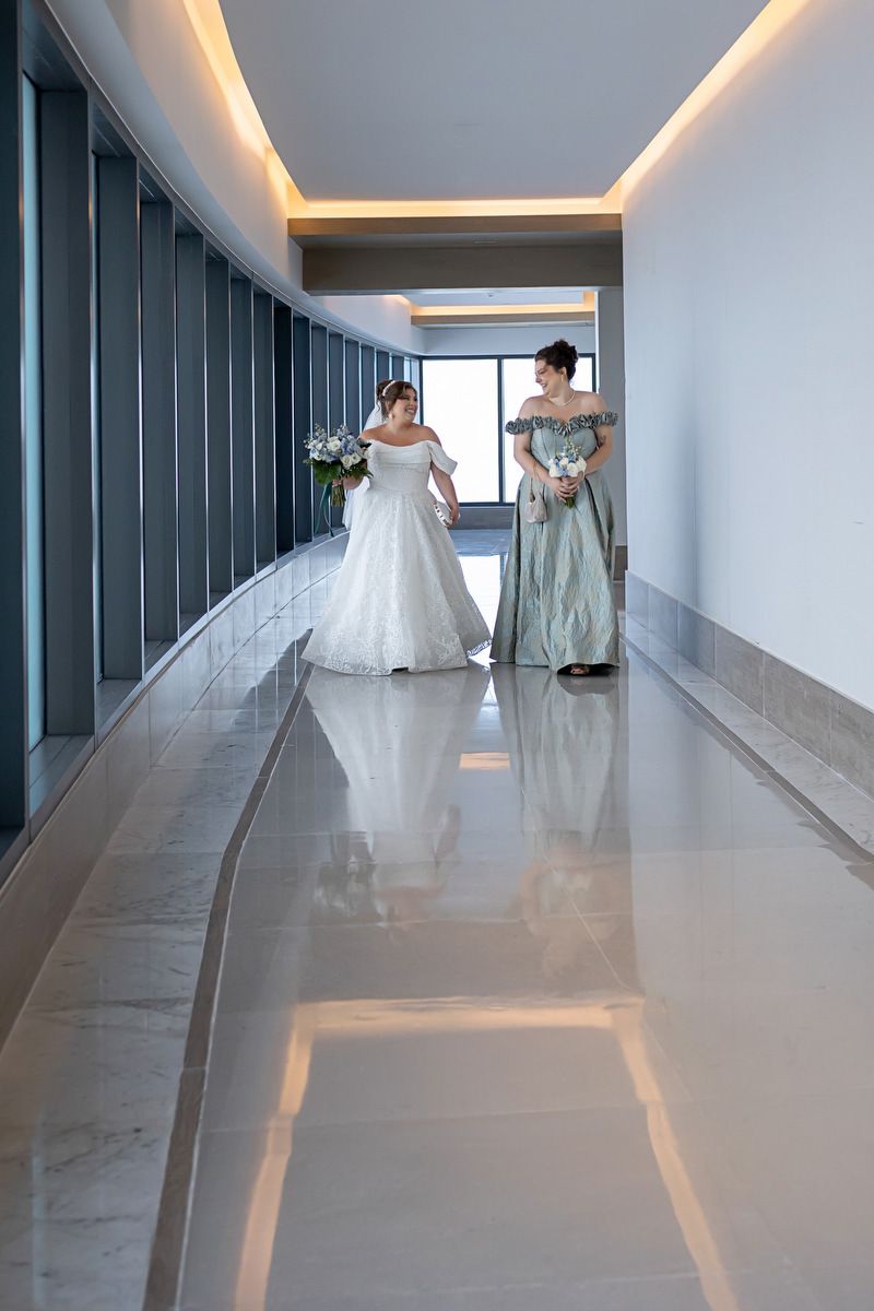 Bride in white off-shoulder gown and bridesmaid in teal dress walking through elegant marble hallway.