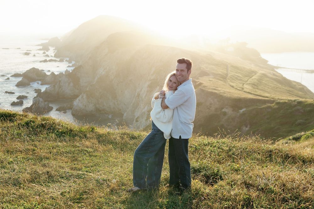 Couple embracing on a coastal cliff at golden hour with ocean and rocky headlands in the background.