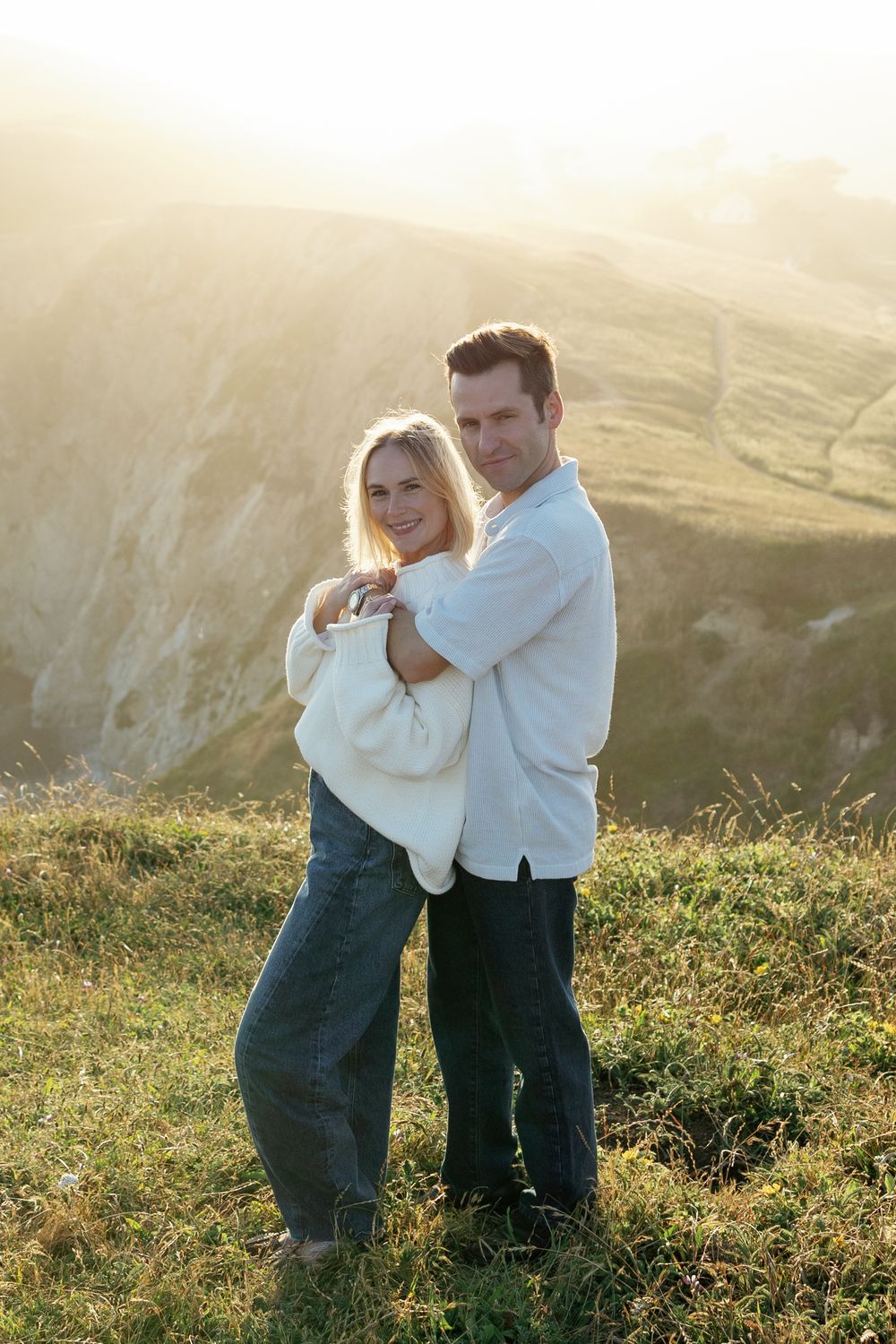 Couple embracing on a sunlit hillside with mountain backdrop, woman in white sweater and jeans smiling warmly.