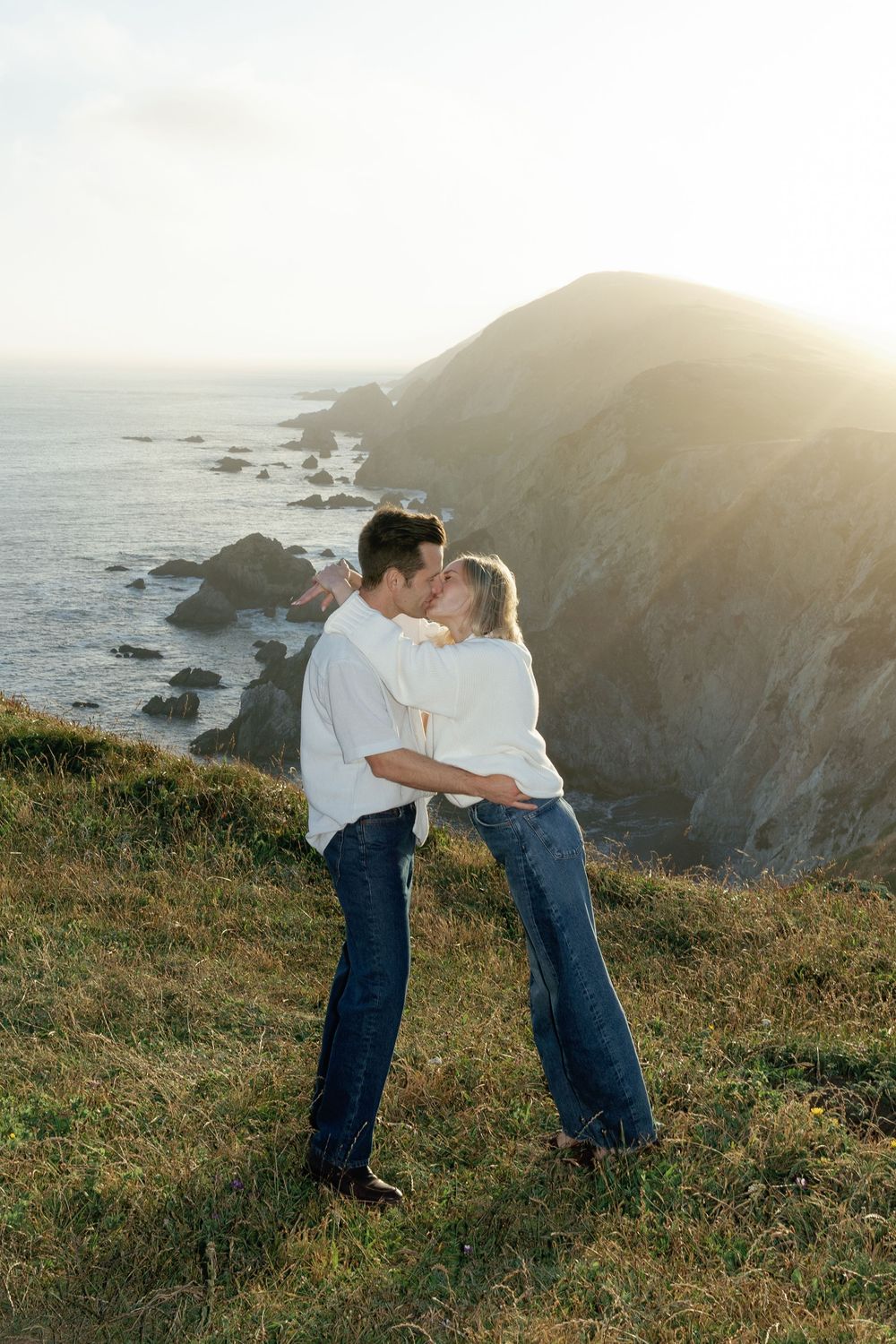 Couple kissing on a coastal cliff at golden hour with ocean and rocky shoreline in the background.