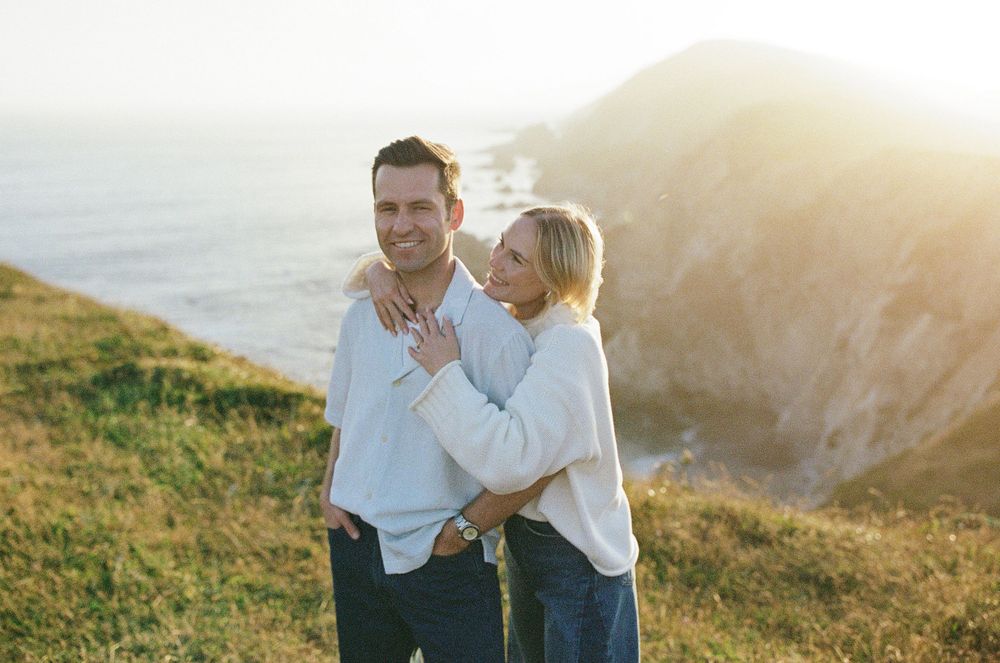 Couple embracing on coastal cliffs at golden hour with ocean and rocky headland in background.