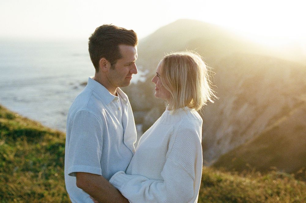Romantic couple gazing at each other on a sunlit coastal hillside, dressed in white with ocean backdrop.