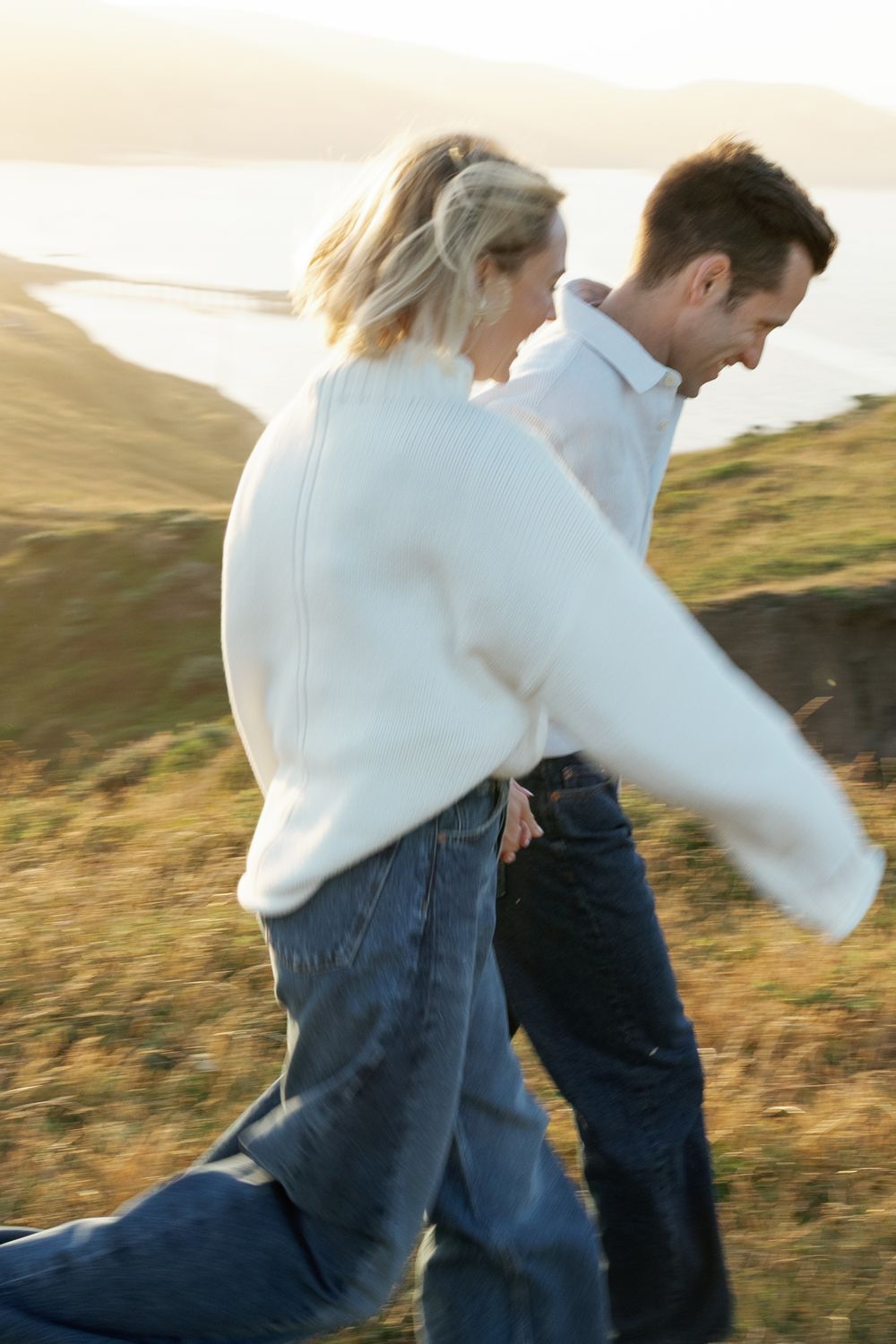 Couple walking hand in hand through golden coastal grasslands with a scenic ocean inlet in the background.