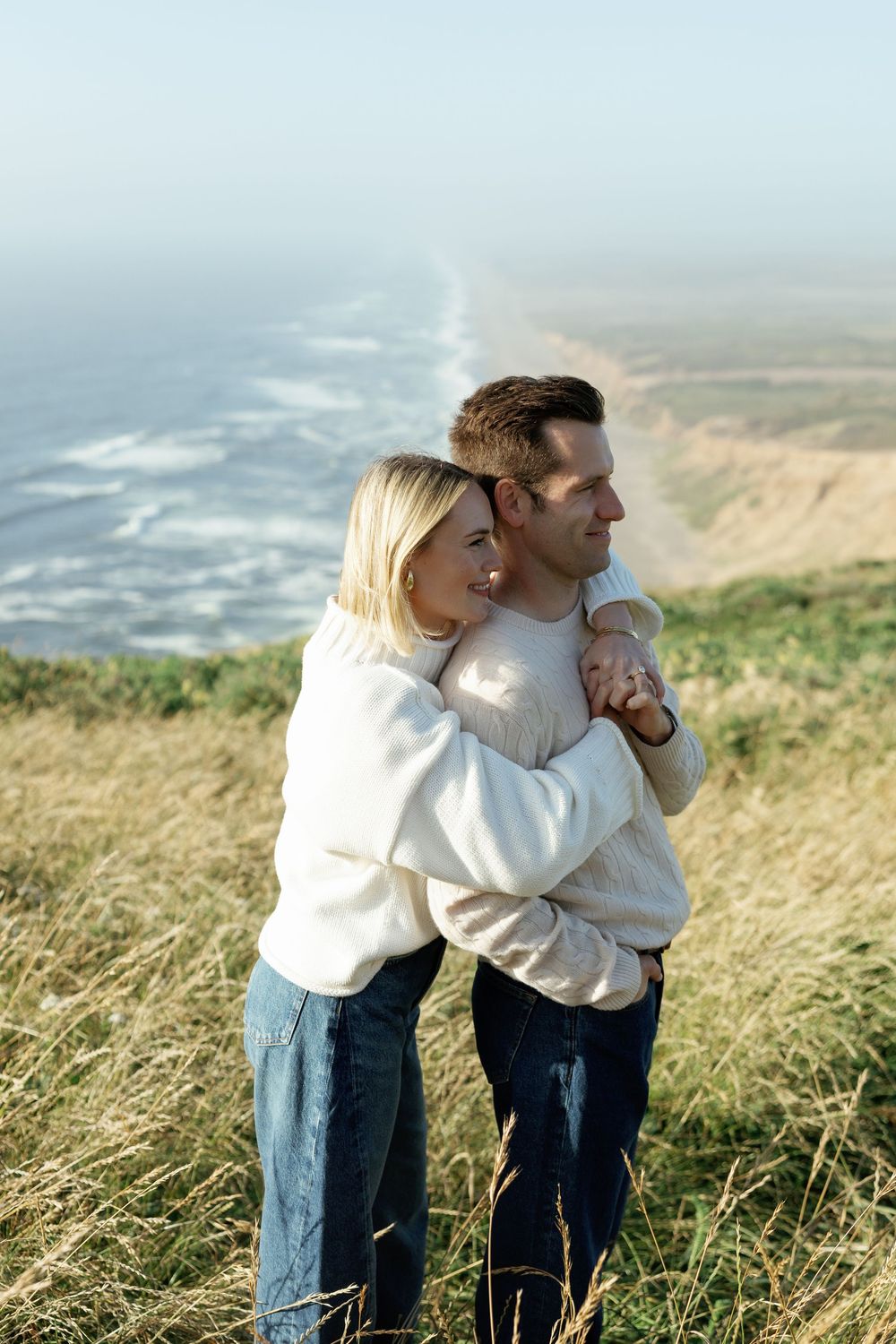 Couple embracing on coastal cliffside meadow, woman hugging man from behind while overlooking ocean shoreline.