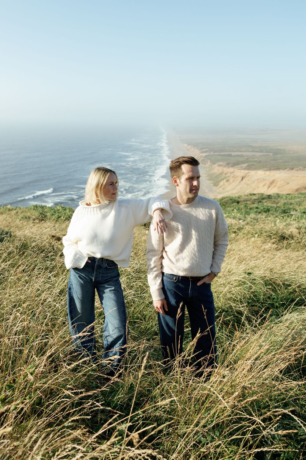 Couple in white sweaters standing on a grassy coastal cliff overlooking the ocean.
