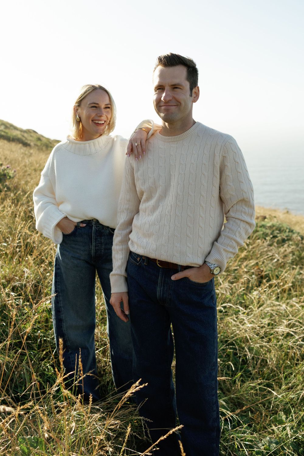 Smiling couple in cream sweaters and jeans standing on a coastal hillside with ocean views.