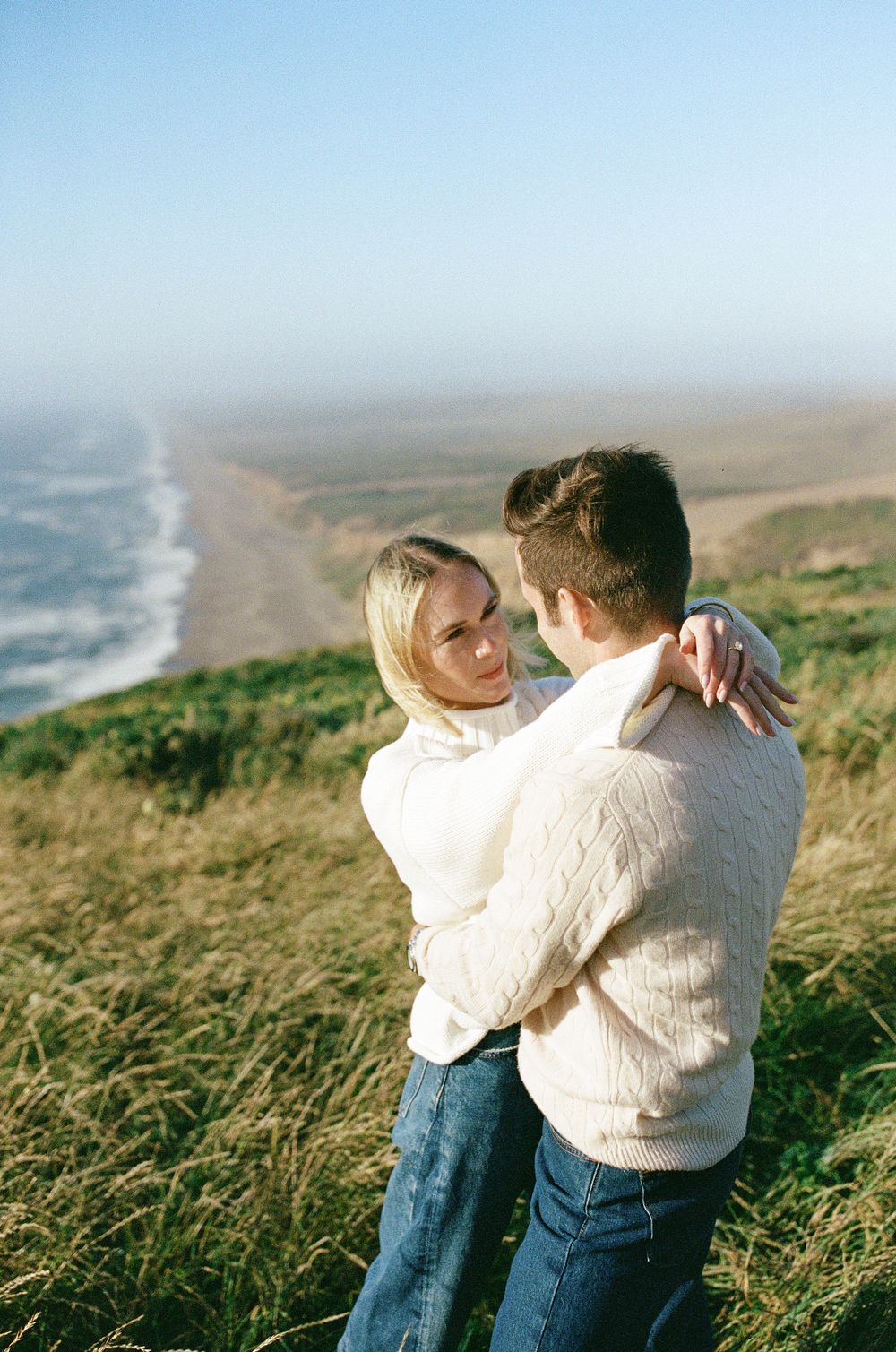 Couple embracing on a grassy coastal bluff with a sweeping view of the beach and ocean behind them.