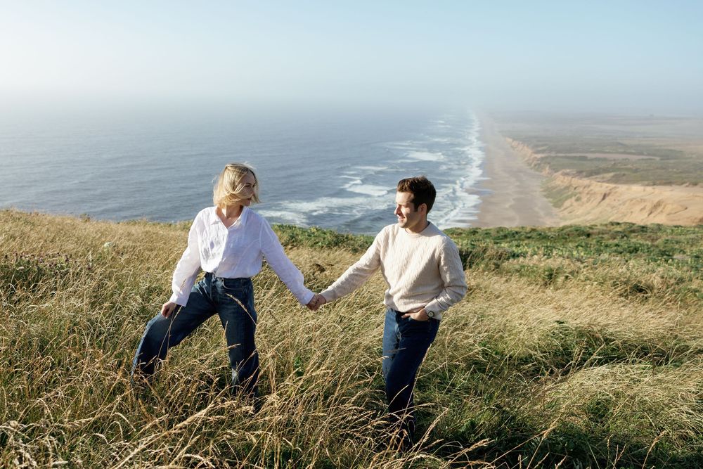 A couple holds hands while walking through golden coastal grass with a scenic ocean beach stretching behind them.