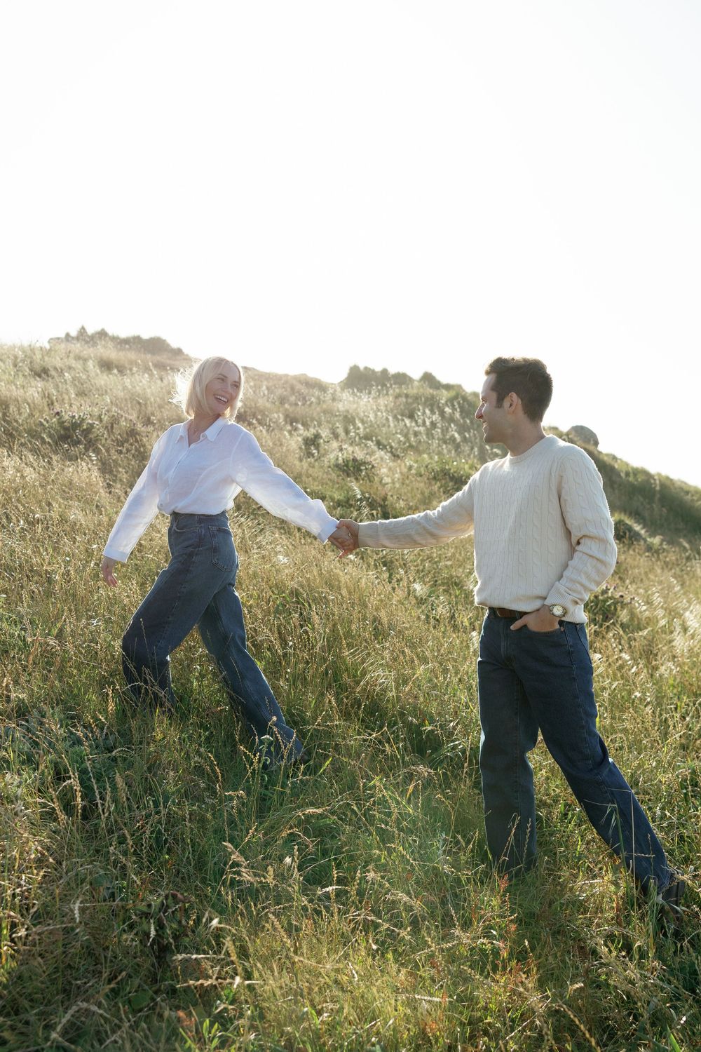 Couple holding hands and walking through tall grass on a sun-drenched hillside meadow.