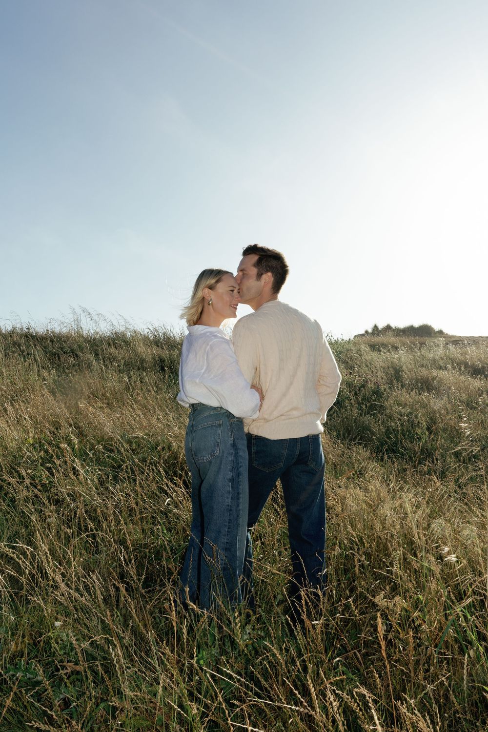 Romantic couple sharing a kiss in tall golden grass field under a bright blue sky.