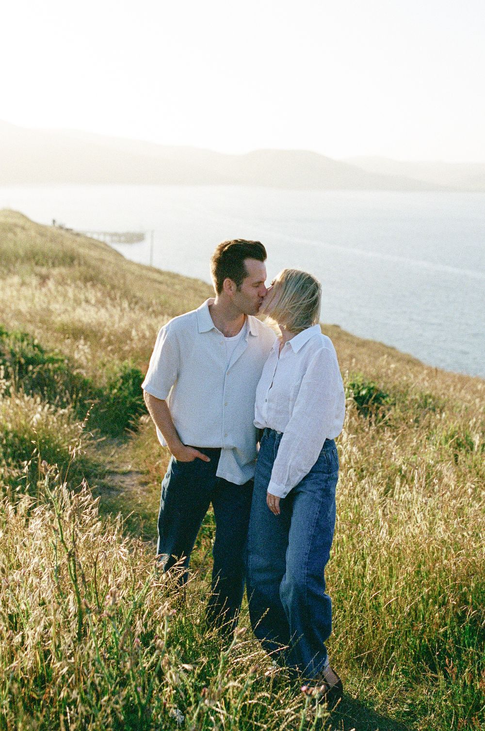 Couple kissing on golden hillside with sparkling ocean and distant hills visible behind them in warm sunlight.