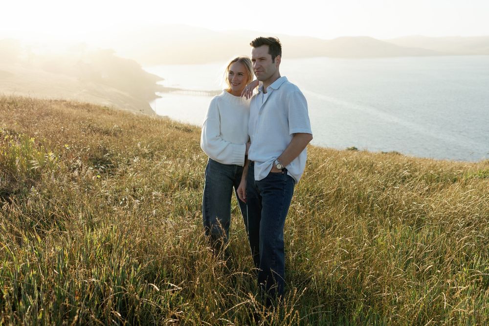 Couple standing together in tall golden grass with sunlit ocean coastline behind them.
